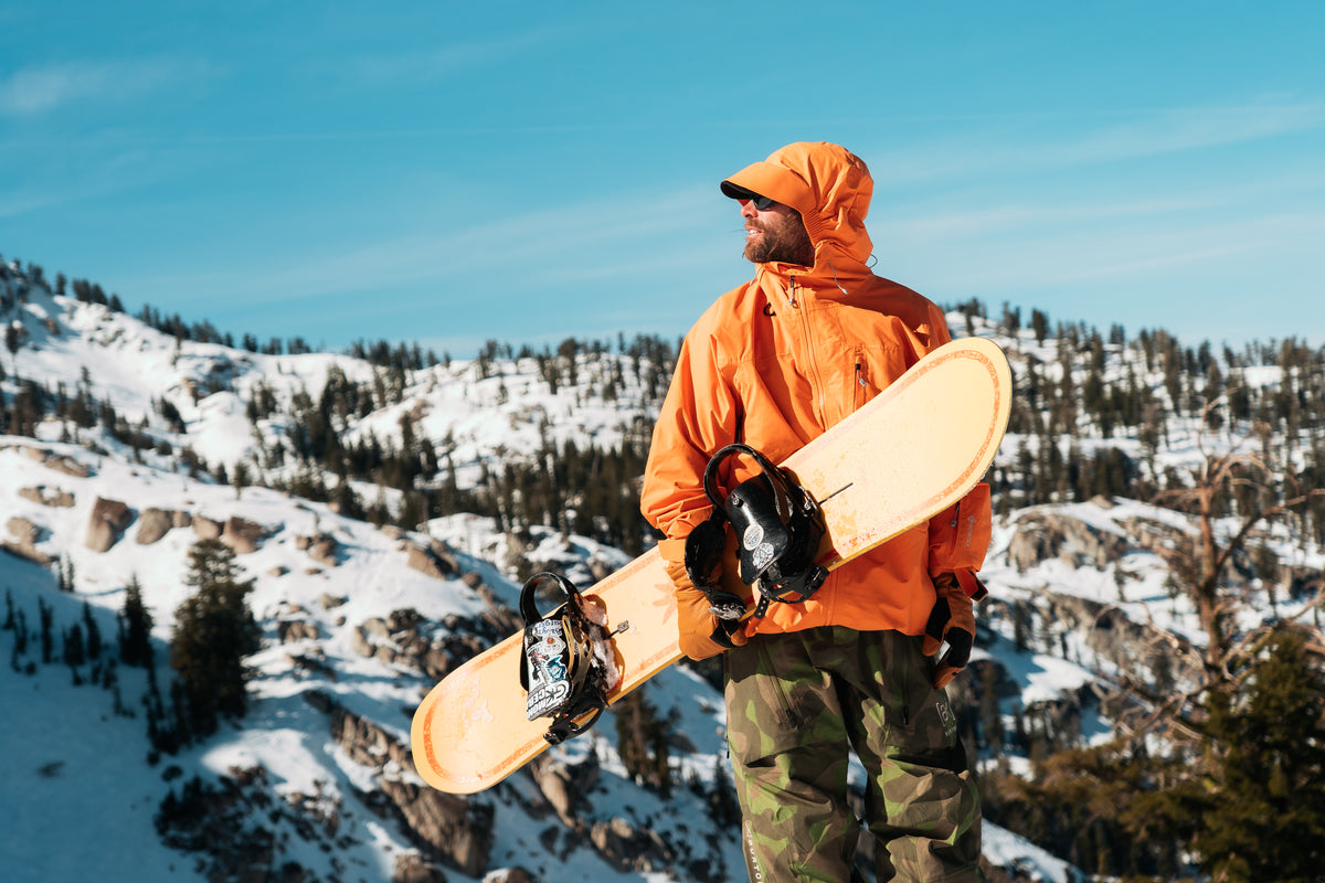 Snowboarder in oranger Jacke mit Snowboard unterm Arm in den Bergen